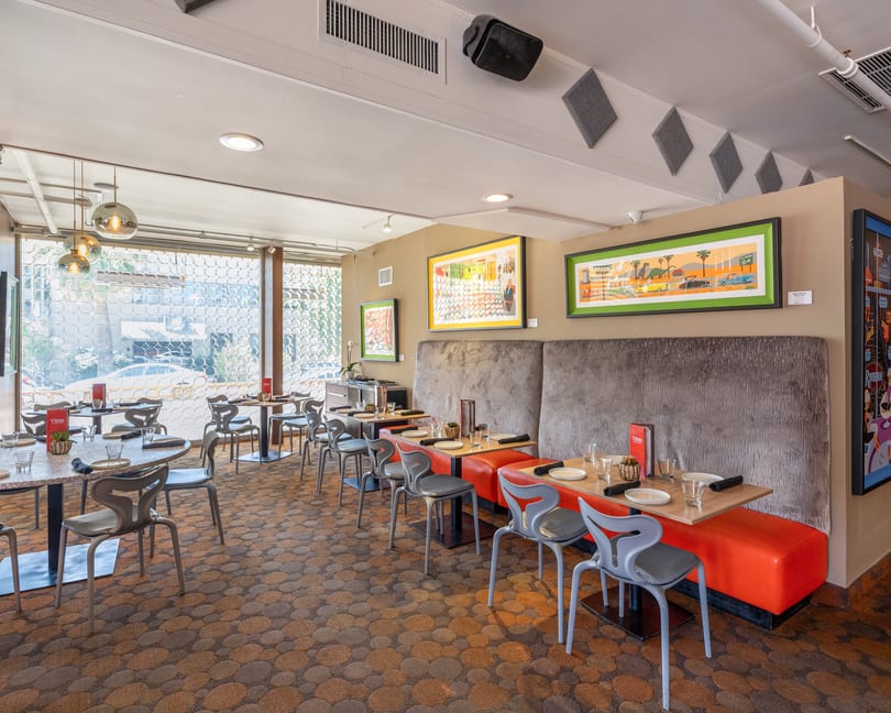 Modern restaurant dining room with orange booths, gray chairs, patterned carpet, and colorful art on the walls.