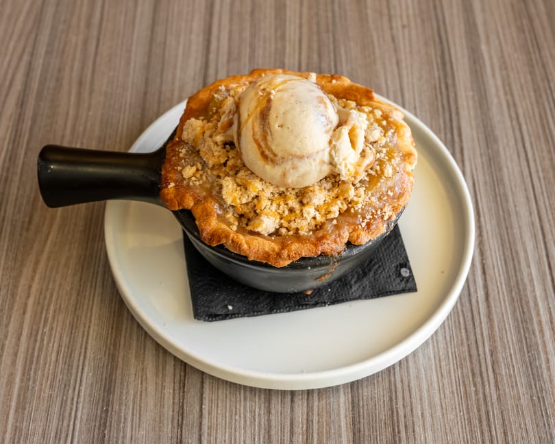 Warm apple crumble pie with a scoop of vanilla ice cream and caramel drizzle in a black skillet.