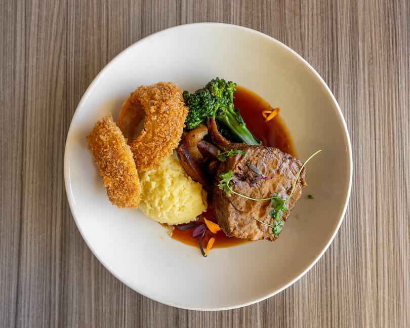 Braised beef with mashed potatoes, onion rings, broccolini, and gravy on a white plate.