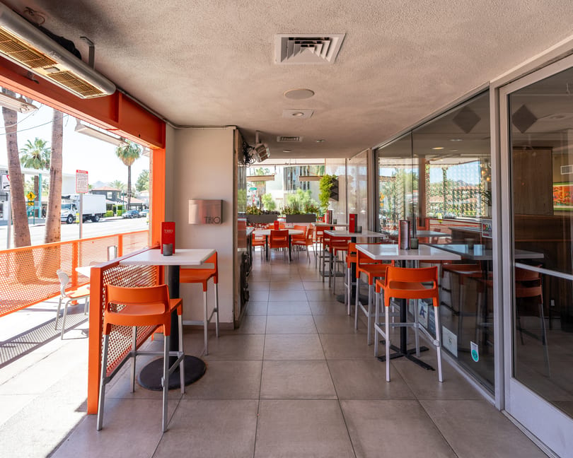Outdoor patio dining area with orange chairs and tables, next to a street with palm trees.