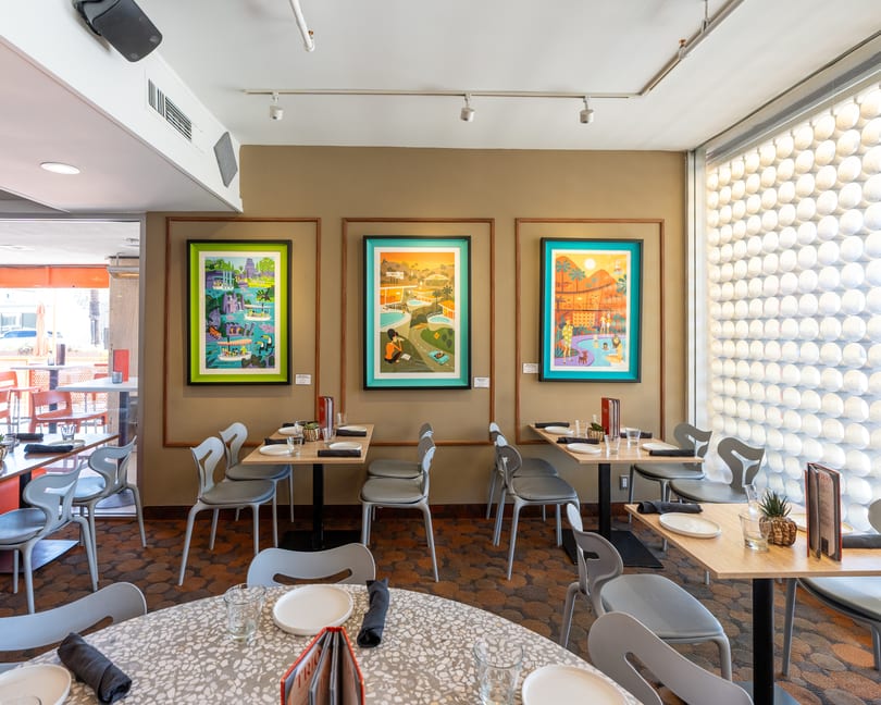 Restaurant dining room with modern tables, gray chairs, and three colorful framed art pieces on a tan wall.