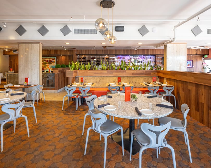 Modern restaurant interior with round tables, gray chairs, orange booths, and a bar with colorful bottles.