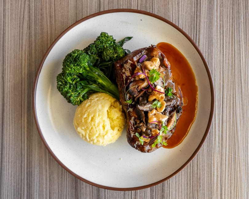 Top-down view of a steak with mushrooms, mashed potatoes, and broccoli on a white plate.