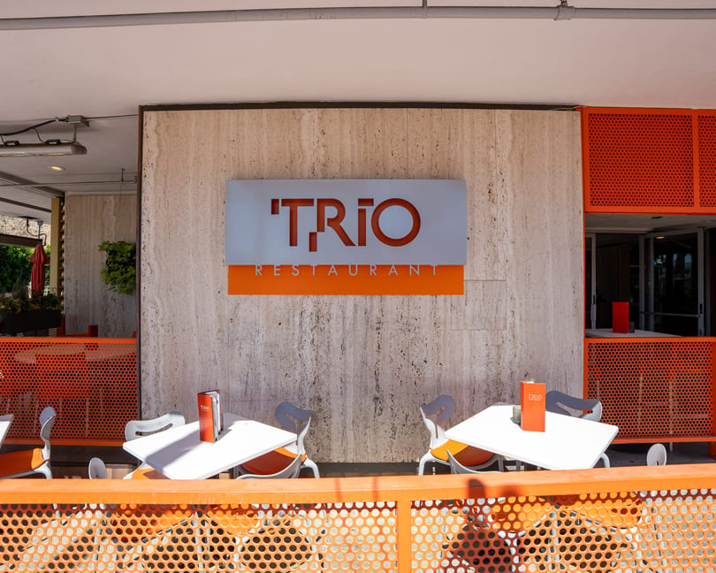The Trio Restaurant sign on a textured wall above an outdoor patio with orange and white tables.