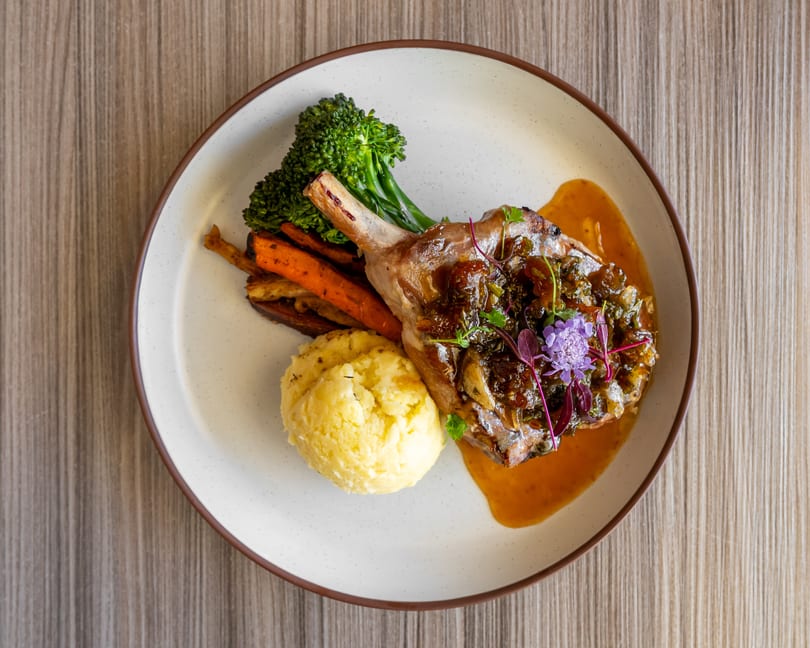 Pork chop with mashed potatoes, roasted carrots, broccolini, and a rich sauce on a white plate.
