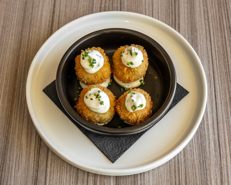 Four golden-brown croquettes topped with white sauce and chives, served in a black bowl on a white plate.