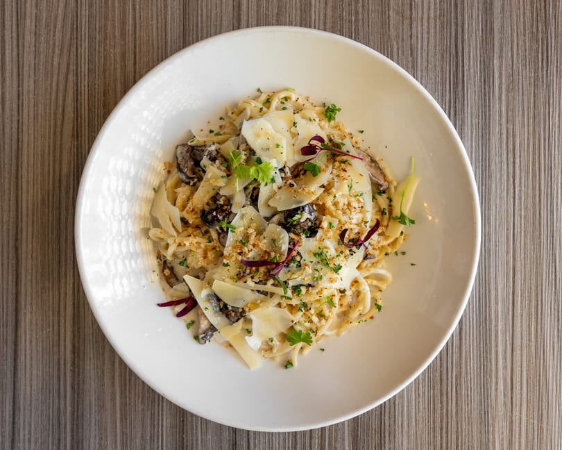 Creamy pasta with mushrooms, shaved Parmesan, breadcrumbs, and fresh herbs in a white bowl.