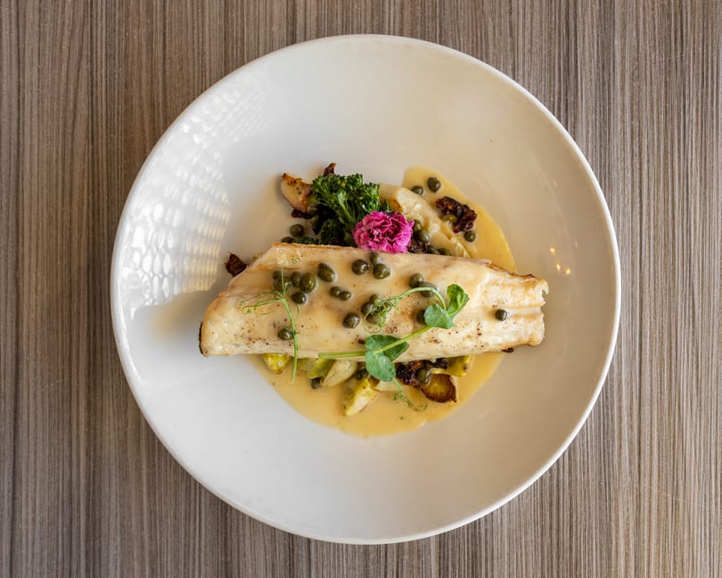 Pan-seared fish with capers, broccolini, sun-dried tomatoes, and a pink edible flower on a white plate.